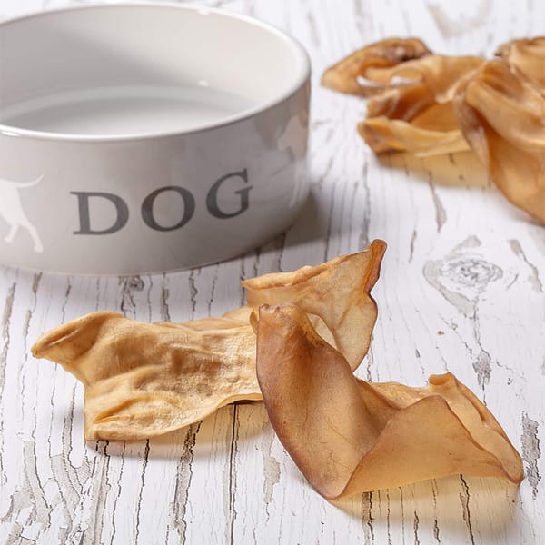 Hollings Cow Ears 3pk Lifestyle 1024 Ceramic Dog Bowl, with Dog written on it, with Dried Cows Ear treats surrounding it